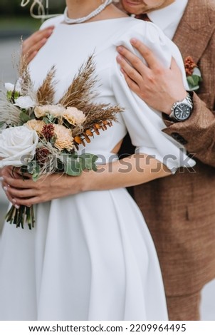 A stylish groom in a brown suit gently hugs the bride in a white dress with a bouquet of wildflowers. Wedding photography, portrait.