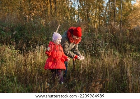A little girl in a red coat walks in nature in an autumn grove with her grandmother. The time of the year is autumn