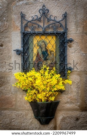 A painting of Mary and Jesus behind a grate with flowers in the foreground, set into a house wall