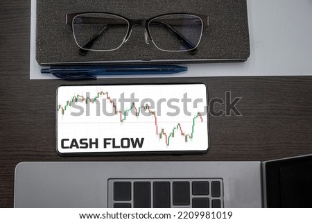 Cash flow text in phone. Top view of chart in the phone on the table near laptop, notepad, and glasses.