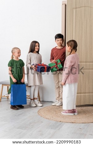 Cheerful interracial kids holding presents near friend and door at home