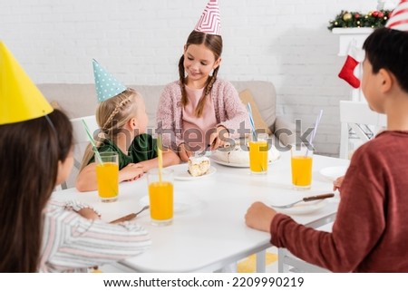 Smiling girl in party cap serving birthday cake to blurred friends near orange juice at home