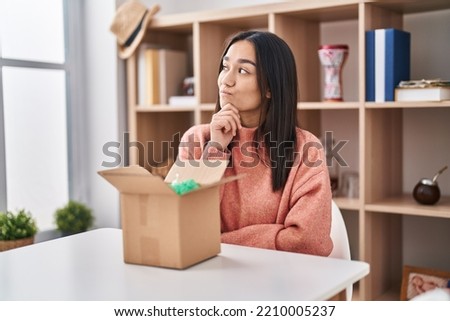 Young south asian woman opening cardboard box thinking concentrated about doubt with finger on chin and looking up wondering 