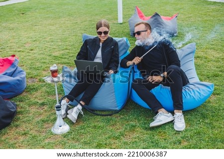 A young woman and a man are sitting in a park on a bean bag and smoking hookah. Happy couple relaxing outdoors with hookah