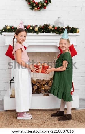 Smiling kids in party caps holding gift and looking at camera near fireplace with christmas stockings at home