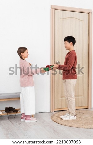 Side view of smiling asian boy giving present to friend near door at home Royalty-Free Stock Photo #2210007053