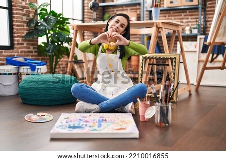 Hispanic woman sitting at art studio painting on canvas smiling in love showing heart symbol and shape with hands. romantic concept. 