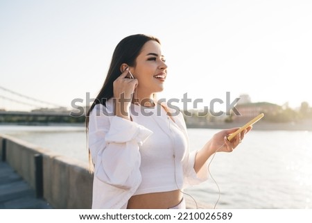 Carefree young female with mobile phone smiling and looking forward while standing on stone embankment and listening to audio book via headphones near river on weekend
