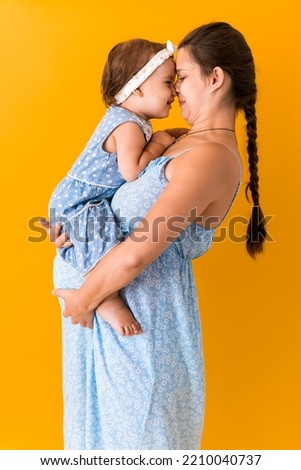 Motherhood, hot summer - portrait young pregnant happy smiling joy mother woman in blue dress holding little baby daughter toddler sibling bare feet in arms looking into eyes kiss on yellow background