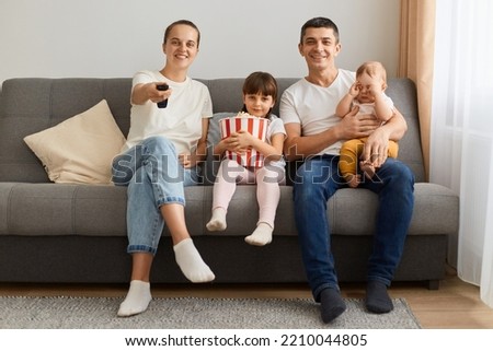 Image of wife and husband wearing casual white t-shirts sitting on cough with their kids, expressing happiness, watching tv, movie or cartoons together.