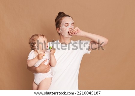Image of sleepy tired woman with bun hairstyle standing with her baby ina hands and with facial mask, doing cosmetic procedures, yawning, covering mouth with hand, isolated over brown background.