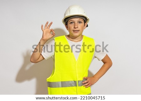 a teenager in a hardhat and a reflective vest shows an ok sign.