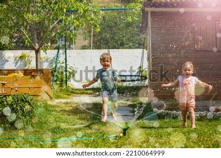 Fun in the garden on hot summer day two children boy and girl run around water sprinkler in the lawn