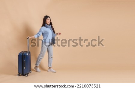 Happy Asian female tourist with luggage in blue sweater and scarf enjoying their winter vacation isolated on beige color background. Excited fashionable 20s woman going on a trip.