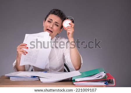 Waist-up portrait of upset girl sitting at the table in office with a pile of documents and a clock, crying because she has no time, isolated on grey background with copy place