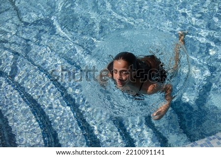 A young teenager girl in a swimsuit, swimming in a blue pool against the background of the sea and sky. The female is diving underwater