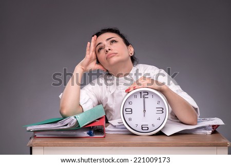 Waist-up portrait of girl sitting at the table in office with a pile of documents and an alarm clock, dreaming about something and lovely looking up, isolated on grey background with copy place