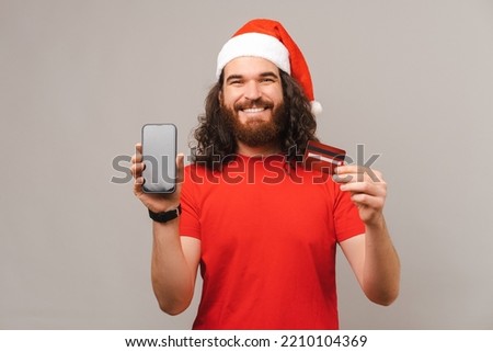 Photo of young happy bearded man wearing santa claus hat and showing credit card and smartphone screen