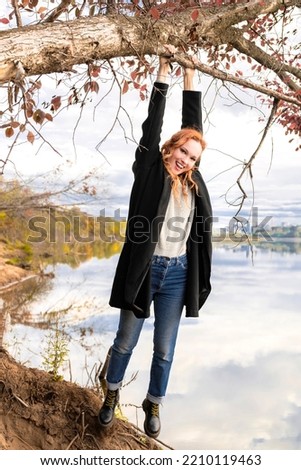 A young woman in a black coat and jeans is hanging and swinging on the branches of a tree on a wild riverbank against the background of an autumn forest.