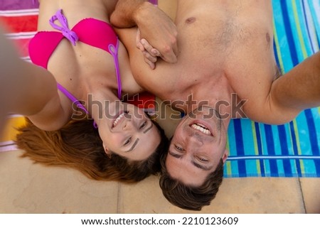 High angle view of a Caucasian couple wearing beachwear lying on towels beside a swimming pool sunbathing and taking a selfie together with a smartphone, out of shot