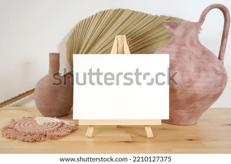 Scandinavian Boho Theme Product Mockup. Table easel and blank card mock up with modern beige ceramic vases with dried palm frond leaf against a white wall background.