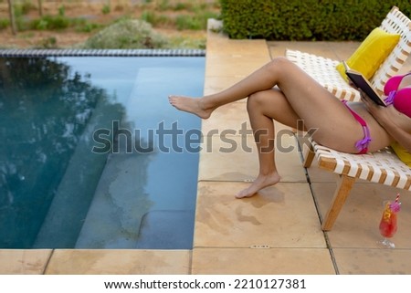 Low section side view of a Caucasian woman wearing beachwear sitting on a chair sunbathing in a garden beside a swimming pool and reading a book