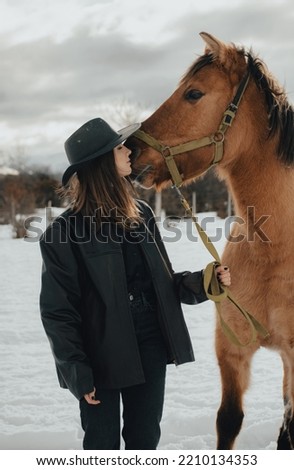 cowgirl kissing her horse in the snow with hat and leather jacket on a cloudy day