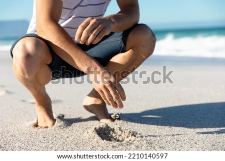 Low section of a Caucasian man squatting on the beach with blue sky and sea in the background, playing with the sand Royalty-Free Stock Photo #2210140597