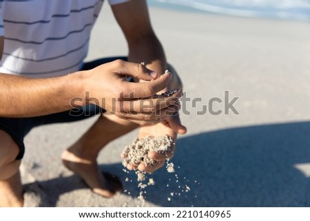 Low section of a Caucasian man squatting on the beach with blue sky and sea in the background, playing with the sand Royalty-Free Stock Photo #2210140965