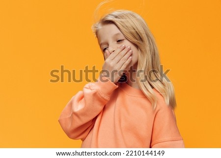 a beautiful girl, blonde, of school age, stands on an orange background in bright clothes and covers her mouth with her hand. Horizontal studio photography with blank space for advertising mockup