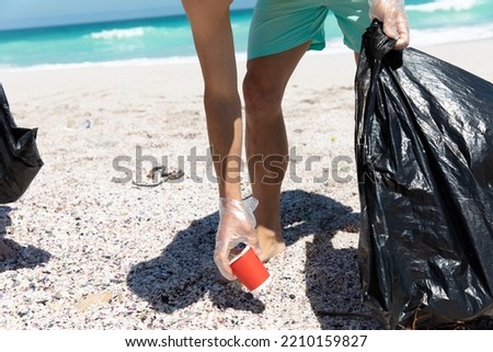 Low section of a Caucasian man on the beach with blue sky and sea in the background, picking up rubbish and putting it into trash bags  Royalty-Free Stock Photo #2210159827