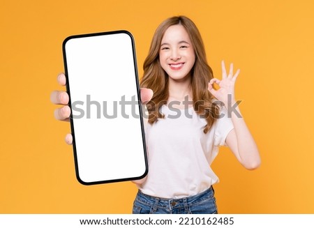 Cheerful beautiful Asian woman holding smartphone and shows ok sign on light yellow background. Royalty-Free Stock Photo #2210162485