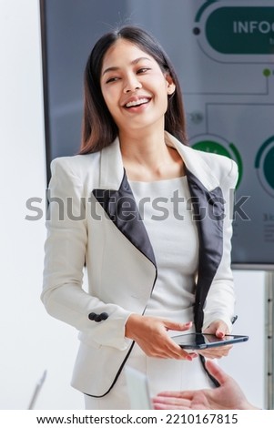 Millennial Asian successful professional businesswoman presenter speaker in white formal suit standing smiling holding tablet while male and female colleagues clapping hands applaud in meeting room. Royalty-Free Stock Photo #2210167815