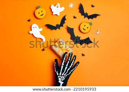 Female hand in funny spooky glove with a gift and Halloween decorations on orange background. Flat lay, top view.