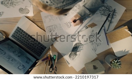 Top view of young woman drawing pencil sketches. Working on a storyboard of a comic in a design studio. Designer work table with a laptop and stationary jar. Royalty-Free Stock Photo #2210197317