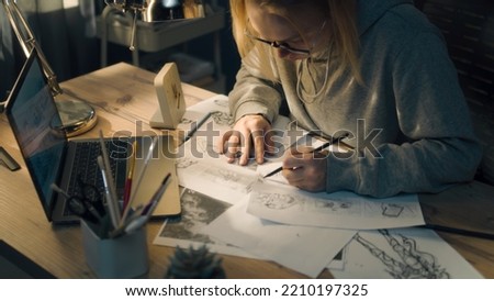 Young creative designer walks towards her work desk in a home based studio. The woman sits on the chair, puts on her glasses and starts sketching with a pencil.