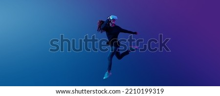 Young Asian woman wearing VR headset with experience playing video game and jumping levitating in the air on futuristic purple cyberpunk neon light banner background. Metaverse technology concept.