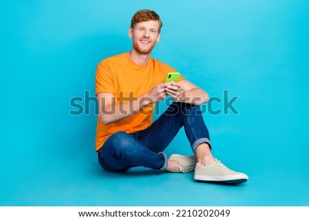 Full length photo of funky positive man wear orange t-shirt sitting floor typing modern device isolated teal color background