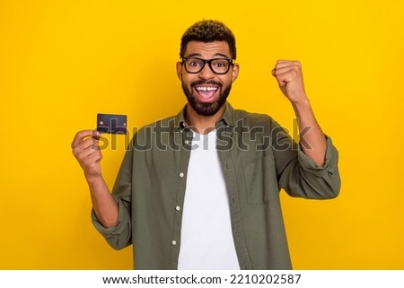 Photo of pretty lucky man wear khaki shirt spectacles rising fist holding credit card isolated yellow color background