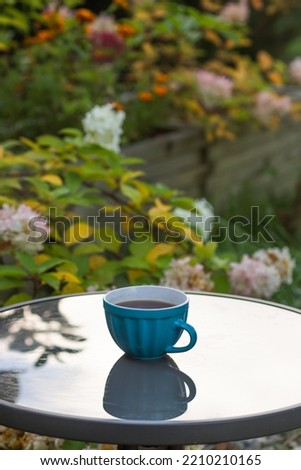 Blue cup of hot coffee on glass round table in the morning garden. Terrace of a rural house with bloom hydrangea flowers