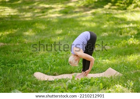 A middle-aged woman practices yoga outdoors, in the Standing Forward Bend pose. Uttanasana.