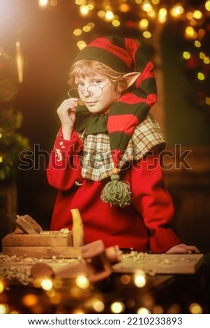 Little cute elf boy makes wooden toys for children for Christmas in the carpentry workshop. Santa Claus workshop. Magic lights flickering around. Christmas tale.