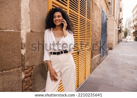 Pleasant young african woman talking on phone walking outdoors during day. Brunette wears singlet, shirt and white casual pants. Concept use, gadgets.