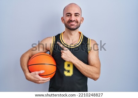 Young bald man with beard wearing basketball uniform holding ball cheerful with a smile on face pointing with hand and finger up to the side with happy and natural expression 