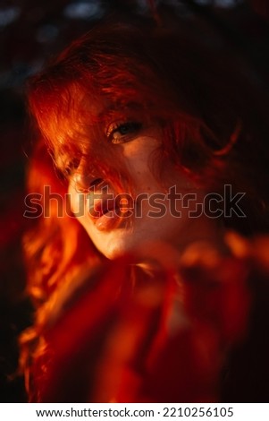Portrait mystical fantasy red-haired woman in red autumn leaves at night. Glamorous lady, red long hair on her face. HAlloween attractive beautiful witch in autumn forest.