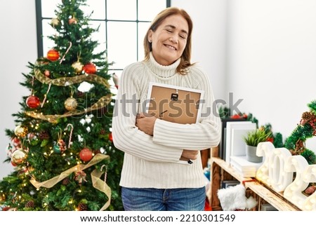 Middle age caucasian woman hugging picture standing by christmas tree at home