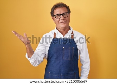 Middle age hispanic man wearing professional cook apron smiling cheerful presenting and pointing with palm of hand looking at the camera. 