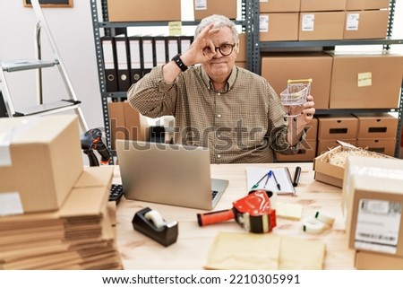 Senior caucasian man working at small business ecommerce holding shopping cart smiling happy doing ok sign with hand on eye looking through fingers 