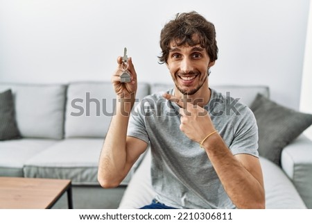 Young hispanic man holding keys of new home sitting on the sofa smiling happy pointing with hand and finger 