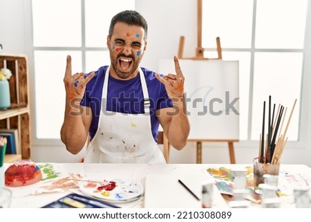 Young hispanic man with beard at art studio with painted face shouting with crazy expression doing rock symbol with hands up. music star. heavy concept. 